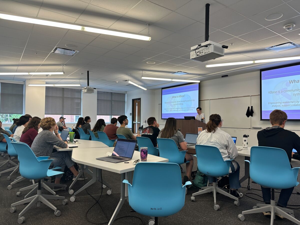 KBase Team Member Zach Crockett presents to a room full of student interns seated in rows in bright blue chairs with their laptops. 