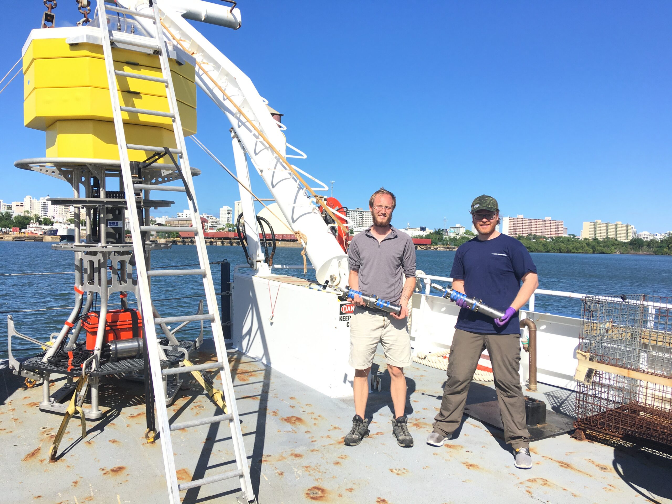 Alvaro Munoz Plominsky, PhD and Logan Peoples, PhD on the Puerto Rico Trench Cruise holding a Pressure Retaining Sampler (PRS). Peoples led the development of the PRS during his PhD. They are standing in front of the Deep Autonomous Profiler (DAP), which was developed by Dr. Christopher Roman and Dr. Steve D'Hondt to sample various depths across the ocean during a single deployment. Photo courtesy of Alvaro Munoz Plominsky.